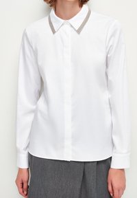 Blusa bianca a maniche lunghe con bottoni nascosti e rifiniture argentate sul colletto, indossata con una gonna grigia plissettata, mostrata dalle spalle ai fianchi.