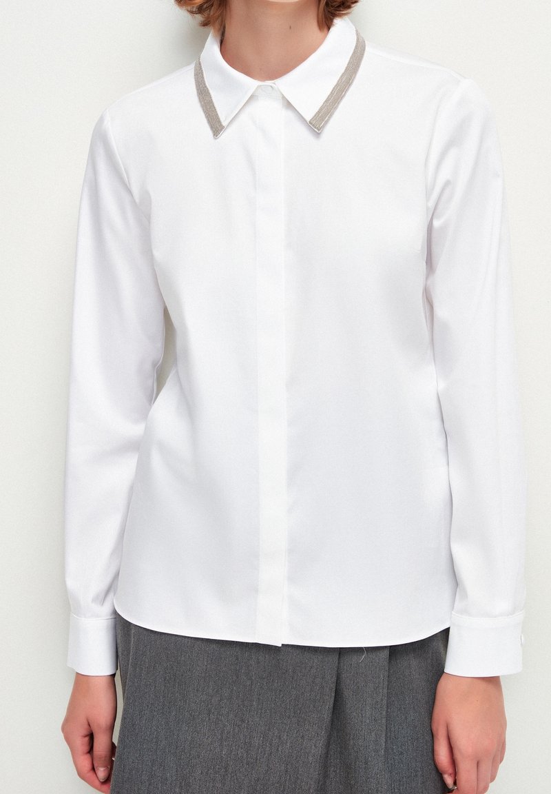 Blusa bianca a maniche lunghe con bottoni nascosti e rifiniture argentate sul colletto, indossata con una gonna grigia plissettata, mostrata dalle spalle ai fianchi.