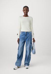 Top a maniche lunghe di crema con maniche attillate, abbinato a jeans cargo blu larghi con molte tasche e dettagli strappati. Tiene in mano una piccola borsa blu.
