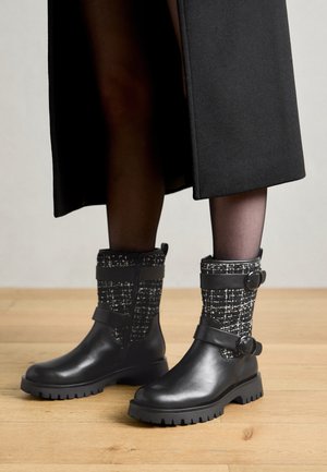 Bottes courtes en cuir noir avec un revers en tissu texturé noir et blanc, dotées de deux brides avec boucles et d'une semelle en caoutchouc robuste.