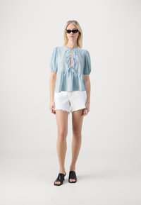 Blusa azzurra chiara con maniche a palloncino, scollatura con lacci e vita arricciata. Abbinata a pantaloni corti bianchi sfilacciati e mule con tacco aperto nere.