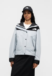 Ανοιχτό μπλε μπουφάν με μαύρες λεπτομέρειες, κουμπιά και κουκούλα. Φορεμένο πάνω από ένα λευκό πουκάμισο, σε συνδυασμό με ένα μαύρο καπέλο με λογότυπο.