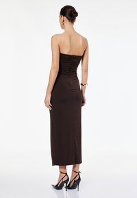 Brązowa, bezrami, dopasowana sukienka z marszczeniem z tyłu, do kolan, w zestawie z czarnymi szpilkami i złotą bransoletką.
