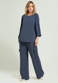 Mujer de pie con una camisa larga de manga larga en azul oscuro y pantalones anchos con bolsillos tipo cargo, combinados con zapatillas plateadas.