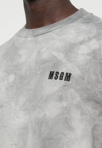 Nahaufnahme einer Person, die ein hellgraues Shirt mit schwarzem, auf der Brust gesticktem MSGM-Logo trägt.