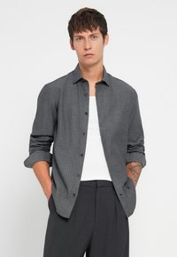 Camisa gris de botones con un sutil patrón de cuadros, mangas largas arremangadas hasta los codos, combinada con una camiseta sin mangas blanca y pantalones oscuros.