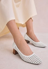 Witte gevlochten hakken met een vierkante teen, voorzien van een gestructureerd patroon en een kleine houten blokhak. Elegant en veelzijdig, passend bij diverse outfits.