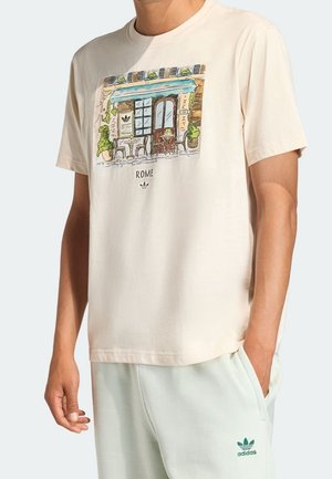 Beżowy t-shirt z kolorowym szkicem fasady kawiarni z napisem Rzym, noszony z jasnymi spodniami Adidas z logo na lewym udzie.