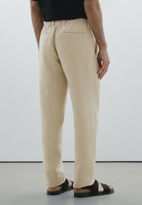 Pantalons en lin beige avec une coupe décontractée, dotés d'une taille avec cordon de serrage et de deux poches arrière, associés à des sandales plates noires.