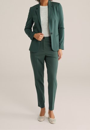 Femme portant un blazer vert ajusté et un pantalon assorti, associé à un haut blanc texturé et des ballerines crème, debout devant un fond uni.