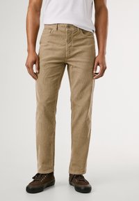 Pantaloni in velluto a coste beige con design a gamba dritta, sottili righe nel tessuto, cinque tasche e chiusura a zip. Indossati con scarpe marrone scuro.