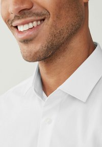 Camisa blanca de botón con cuello puntiagudo y textura suave, que presenta un solo botón en el cuello y un diseño ajustado.