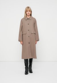 Beige, knee-length coat made of smooth fabric, featuring a large collar, front buttons, and two patch pockets. Worn with black boots.