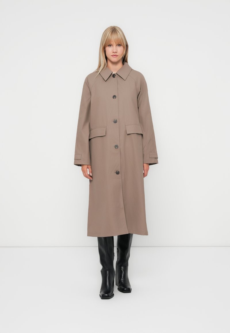 Beige, knee-length coat made of smooth fabric, featuring a large collar, front buttons, and two patch pockets. Worn with black boots.