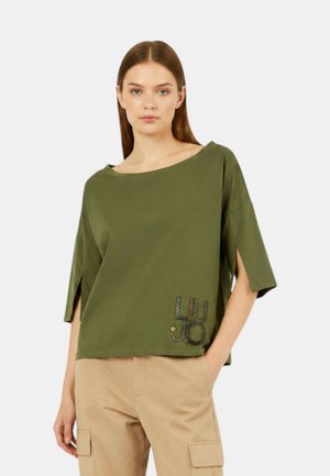 Donna che indossa un top largo verde oliva con spacchi laterali e pantaloni cargo beige con le mani nelle tasche su sfondo bianco.