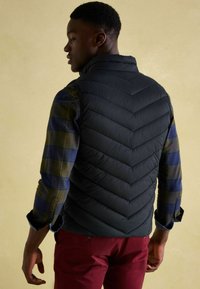 Junger Mann, der seitlich blickt, trägt eine schwarze gesteppte Weste über einem grün-blau karierten Hemd und roten Hosen vor einem beigen Hintergrund.