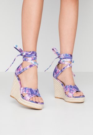 Pieds portant des sandales compensées à lanières autour de la cheville, tie-dye violet et rose, avec talons plateforme tissés sur un fond neutre.