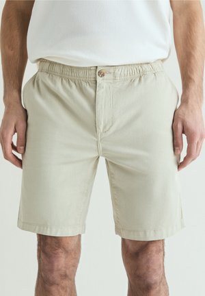 Hombre con pantalones cortos beige con cintura elástica y botones delanteros y camisa blanca, manos descansando a los lados sobre un fondo liso.