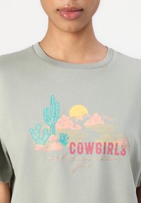 Light green cotton t-shirt featuring a graphic with cacti, mountains, a sun, and text "COWGIRLS just wanna have fun" in pink and pastel colors.