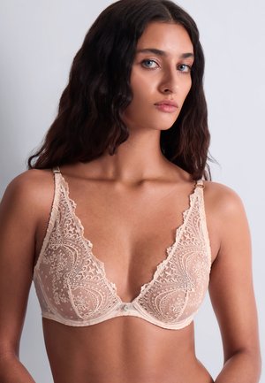Femme aux longs cheveux foncés portant un soutien-gorge en dentelle beige clair à motifs floraux, posant devant un fond uni gris clair.