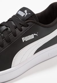 Černá sneaker Puma s bílým pruhem, černými tkaničkami a logem Puma na boku a jazyku, zobrazená zblízka.