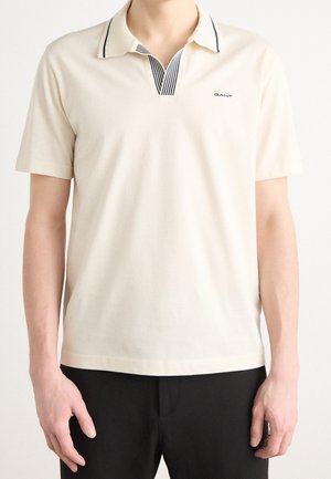 Homme portant un polo crème à manches courtes avec un col rayé noir et le logo GANT sur la poitrine, associé à un pantalon noir.