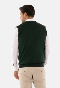 Maglione senza maniche di un verde scuro, realizzato in morbido materiale a maglia, mostrato di schiena, abbinato a una camicia bianca con bottoni e pantaloni chiari.