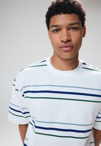 T-shirt bianca di cotone con righe orizzontali in tonalità di blu e verde. Maniche corte, collo rotondo, tessuto morbido e vestibilità casual.