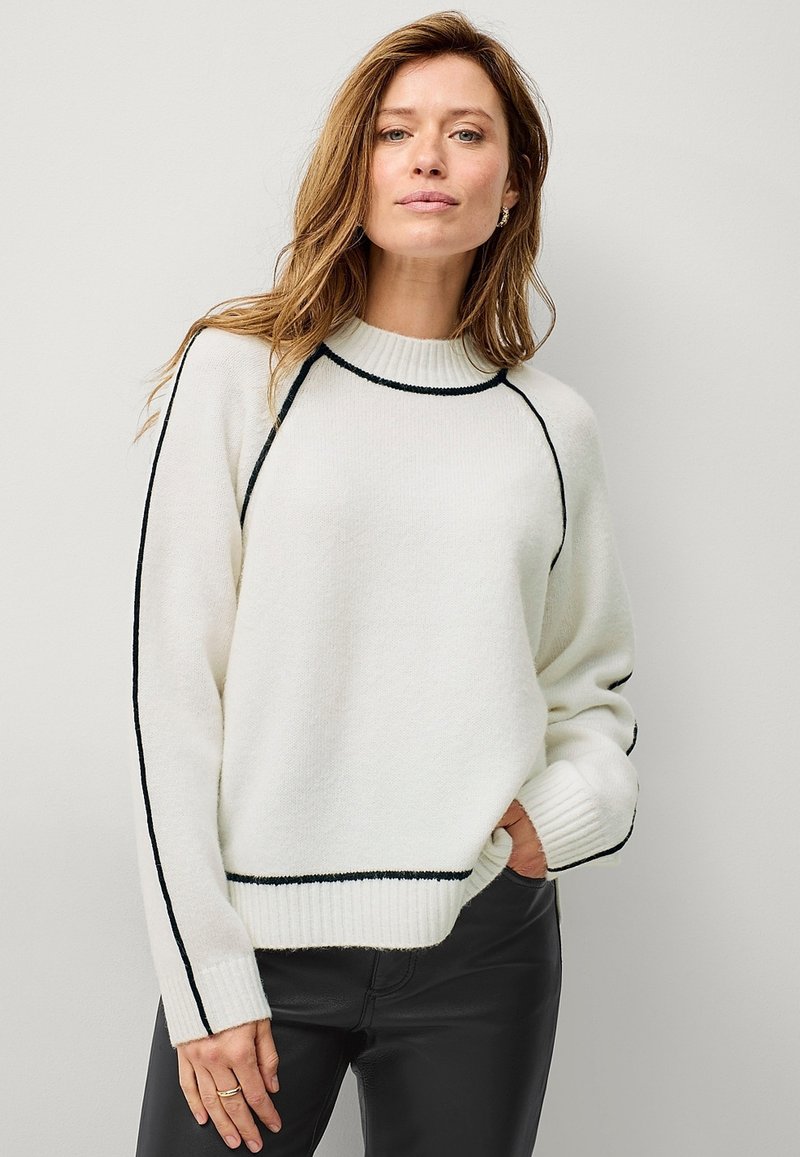 Maglione bianco lavorato a maglia con un collo alto, rifiniture nere lungo le spalle e i polsini, e maniche lunghe, abbinato a pantaloni neri.