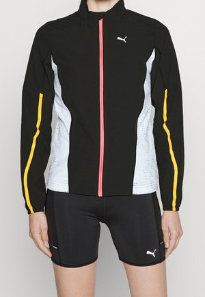 Person trägt eine schwarze Puma-Jacke mit weißen Seitenteilen, gelben Streifen an den Ärmeln und einem leuchtend pinken Reißverschluss, kombiniert mit schwarzen Puma-Shorts.