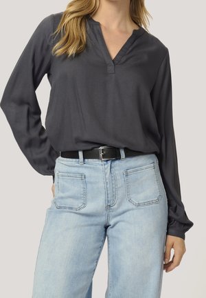 Frau trägt ein dunkelgraues Langarmshirt mit V-Ausschnitt, das in hellblaue High-Waist-Jeans mit vorderen aufgesetzten Taschen und einem schwarzen Gürtel gesteckt ist.