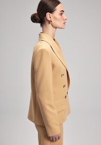 Beige dvouřadý blejzr s přiléhavým střihem, strukturálními rameny a zlatými knoflíky, doplněný o odpovídající kalhoty. Hladká textura látky.