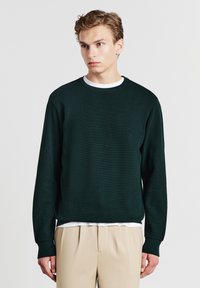 Maglione a maglia di un verde scuro e strutturato con scollatura rotonda. Indossato sopra una camicia bianca, abbinato a pantaloni beige. Maniche lunghe e vestibilità comoda.