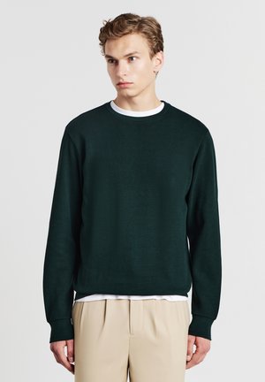 Dunkelgrüner, strukturierten Strickpullover mit Rundhalsausschnitt. Über einem weißen Hemd getragen, kombiniert mit beigen Hosen. Lange Ärmel und lockere Passform.