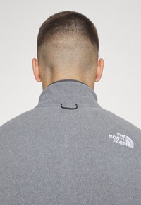 Gray fleece pullover with a high collar, featuring a small black loop at the back and a white logo on the right shoulder.