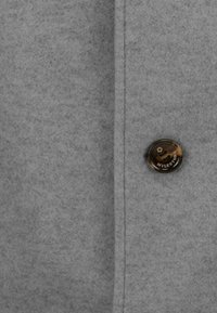 Grauer, weicher Stoff mit glatter Textur; verfügt über einen runden, dunklen Knopf mit marmoriertem Design und eingraviertem Text, der sich auf der linken Seite befindet.