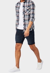 Hombre caminando con una camisa de cuadros arremangada sobre una camiseta blanca, pantalones cortos de mezclilla oscuros y zapatillas blancas, contra un fondo liso.