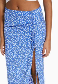 Modrá květinová zavinovací sukni s nařaseným pasem, se štěrbinkou a detaily s tkaničkami. Materiál vypadá lehce a má jemnou texturu.