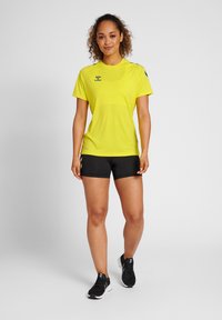 Gul atletisk t-shirt med korte ærmer, sorte shorts og sorte sportssko. Har logo på brystet og kontrastfarvet accenter på ærmerne.