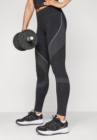 Fekete, magas derekú leggings, texturált szürke csíkokkal, fekete sportcipővel párosítva, súlyzót tartva egy edzőtermi környezetben.