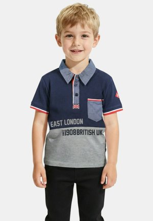 Jeune garçon blond souriant, portant un polo bleu marine et gris avec les inscriptions East London et British UK, ainsi qu'un pantalon noir, debout devant un fond blanc.