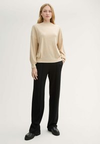 Beżowy, oversize sweter z długim rękawem i wysokim kołnierzem, zestawiony z czarnymi, wysokim stanem, dopasowanymi spodniami oraz czarnymi butami na grubej podeszwie.
