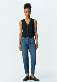 Chaleco negro sin mangas con escote en V, que cuenta con seis botones y bolsillos laterales; combinado con vaqueros de cintura alta azules y mocasines negros.