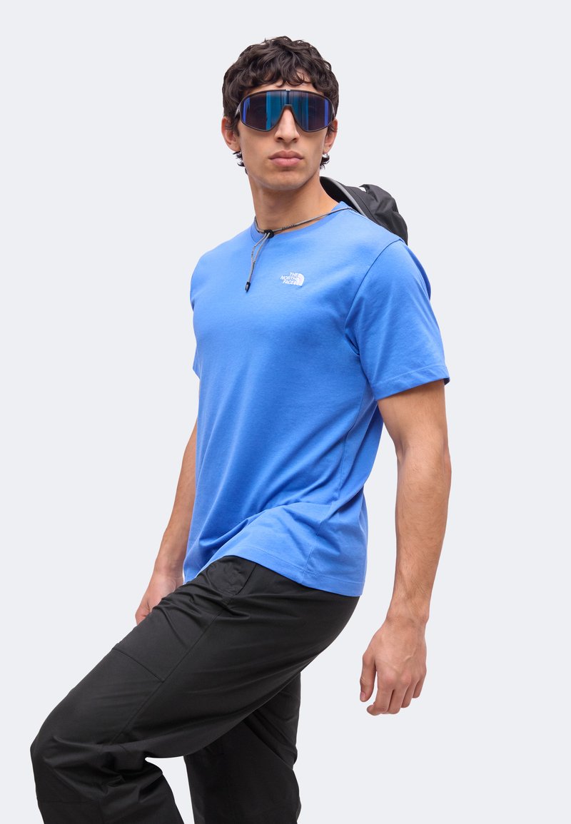 Joven con camiseta azul de North Face, pantalones negros, grandes gafas de sol reflectantes y una correa de mochila sobre un hombro, de pie frente a fondo blanco.