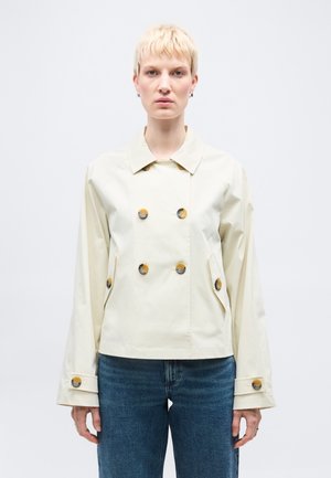 Osoba o krótkich blond włosach, ubrana w kremową dwurzędową kurtkę z brązowymi guzikami i niebieskie dżinsy, zwrócona twarzą do przodu.