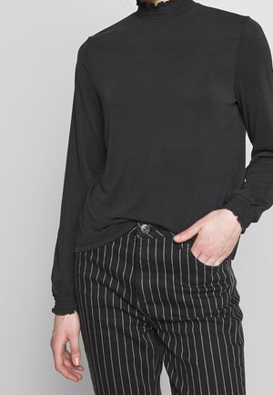 Person trägt ein schwarzes Langarmoberteil mit gerafftem Kragen und Manschetten, kombiniert mit schwarzen Hosen mit Nadelstreifen, eine Hand in der Tasche.