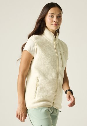 Young woman wearing a cream-colored fleece vest over a short-sleeve shirt and light green pants, standing against a plain background.