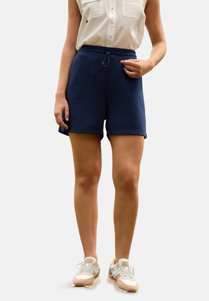 Mujer con pantalones cortos de cintura elástica azul marino, blusa blanca sin mangas con textura y zapatos deportivos beige, de pie frente a un fondo liso.