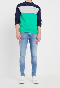 Laivansininen, harmaa ja vihreä puuvillaneule pitkillä hihoilla; yhdistetty kevyisiin sinisiin slim-fit farkkuihin ja valkoisiin urheilukenkihin.