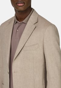 Blazer beige de espiga con solapas en pico y un único bolsillo en el pecho, superpuesto a una camiseta polo color malva. Textura suave, corte entallado.
